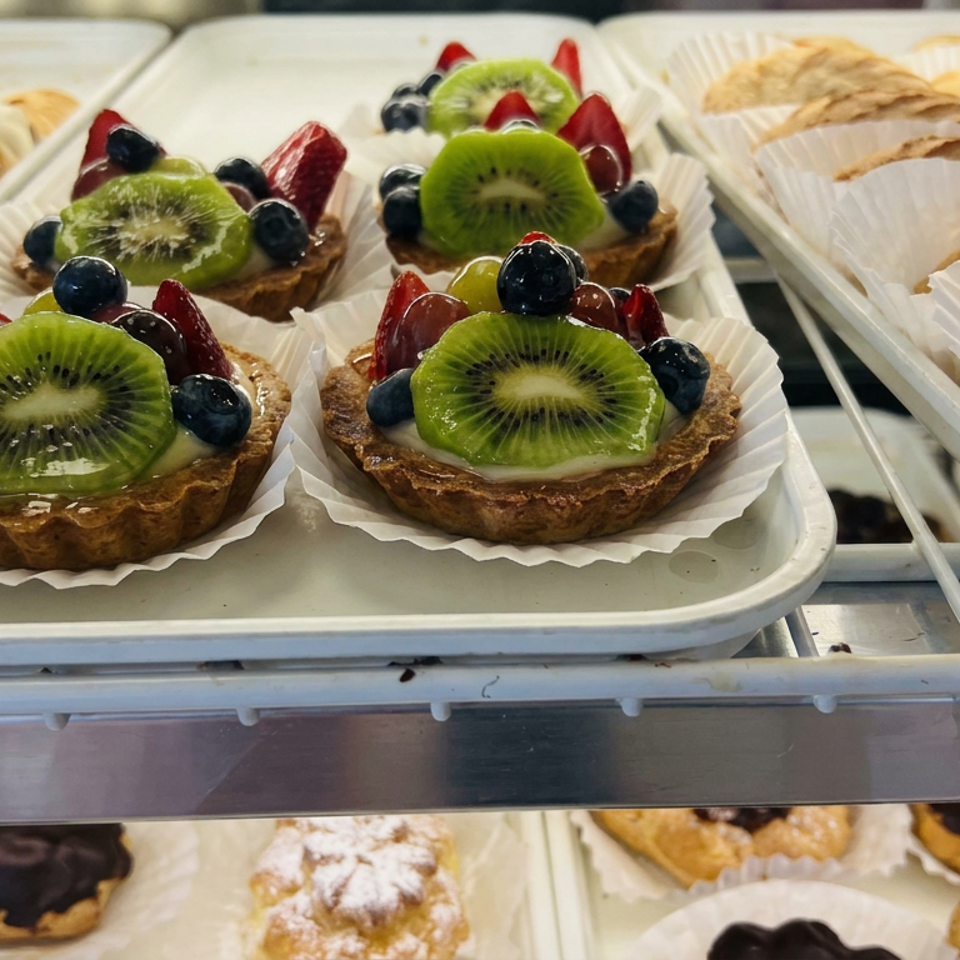 Fruit tarts topped with kiwi, strawberries, and blueberries displayed alongside assorted pastries in a bakery case.