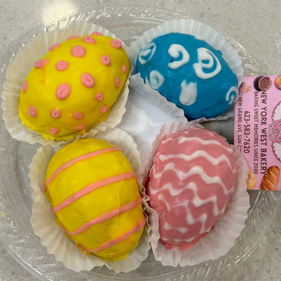 Four colorful frosted pastries sit in paper cups on a clear plate beside a New York West Bakery business card.
